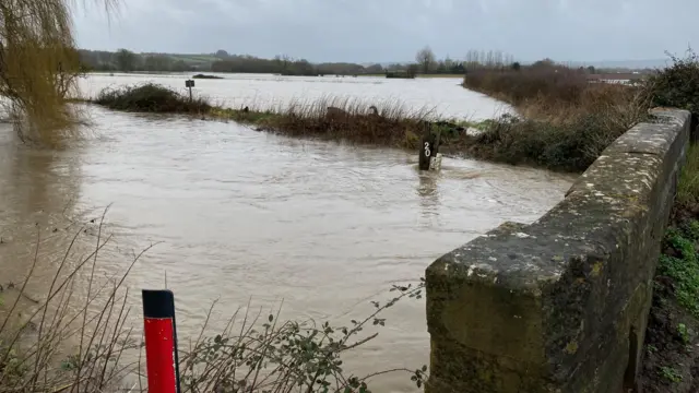 A very swollen river passes under a stone bridge. A marker shows the water has nearly hit 2m. Fields in the background are flooded.
