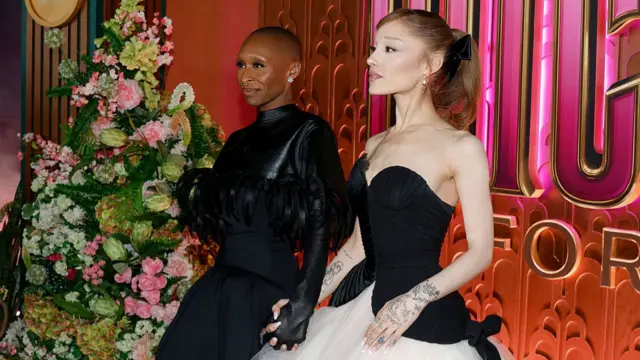Cynthia Erivo, wearing a black feathered dress, holds hands with Ariana Grande, wearing a black and white strapless dress, in front of a sign just seen reading Wicked: For Good - they are both looking off camera