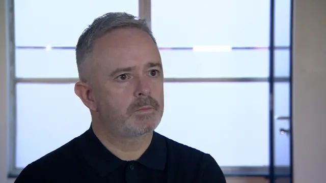 Paul Bowen, a man with short, greying hair and stubble, wearing a black polo shirt during a TV interview.