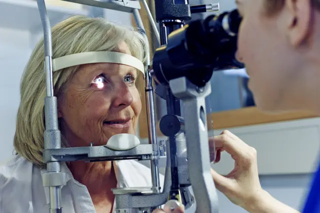 An older woman having an eye test. She has blue eyes and straight, blonde hair to her shoulders and she is wearing a light-coloured shirt. A healthworker is examining her eye with testing equipment.