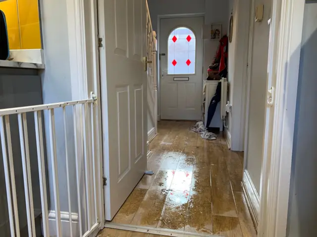 A hallway in a house, taken from the kitchen and through to the front door. The brown laminate flooring is wet, and a wet towel can be seen near the front door.