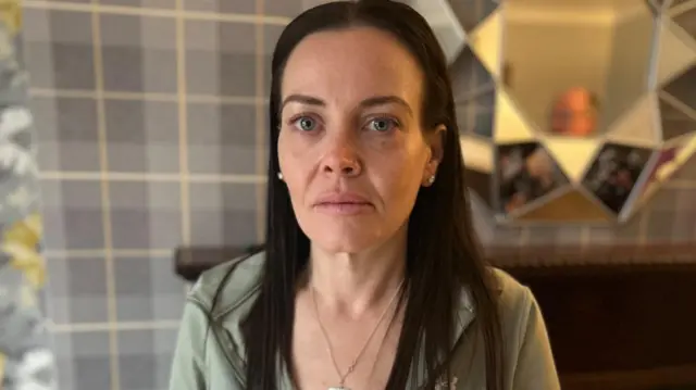 A woman with long brown hair looks into the camera. She is wearing a green zipped up hoody and is standing in front of grey and yellow wallpaper and a mirror.