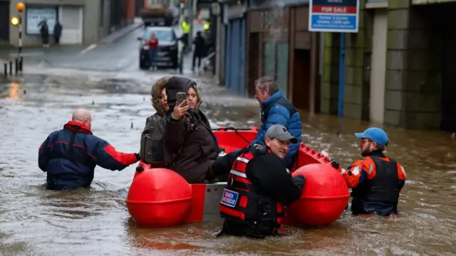 Three people sit in a boat being pushed along by a rescue team in a flooded street