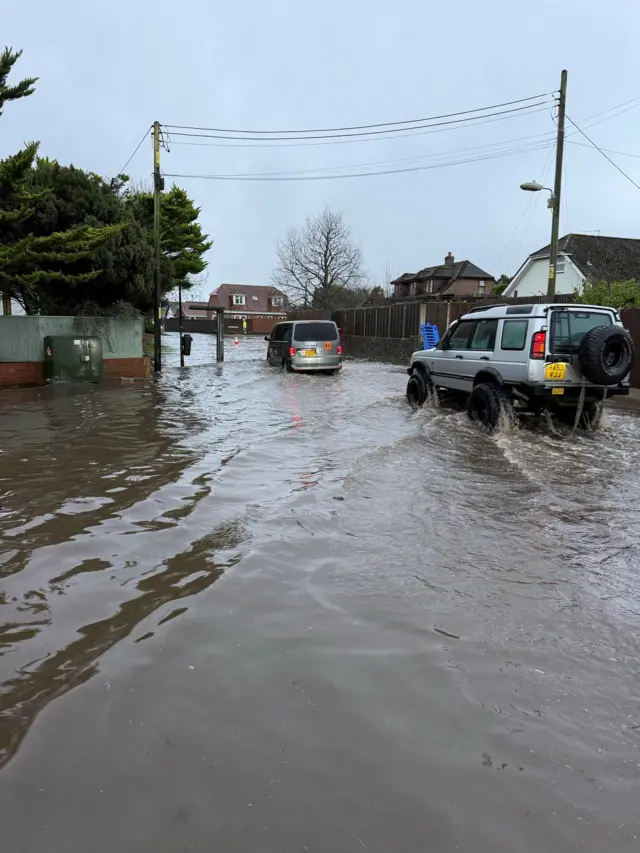 A flooded Eastfield Lane, Junction to Hightown road, in Ringwood. There are cars driving through the water.