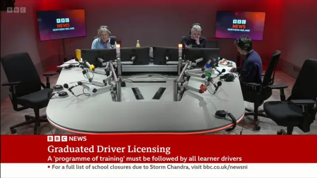 An image showing inside the BBC Radio Ulster studio - we can see a large grey table, on which there is a number of monitors and microphones. Three people are around the table, on the left a woman with headphones on, in the middle a man and on the right another woman. In the background on the wall are monitors with the BBC News NI logo