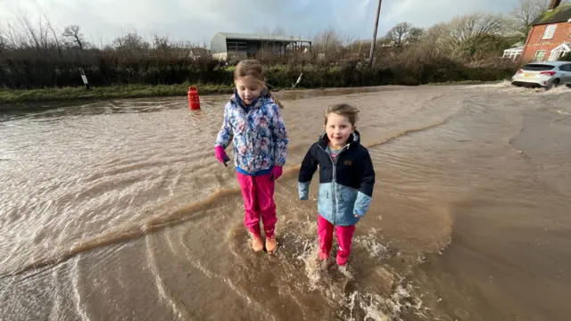 Two young girls pictured wearing wellies and waterproofs while standing on flooded water