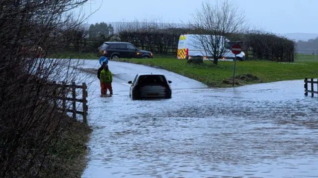 A car driving through floodwater away from the camera. In the foreground is a large amount of water. There are people standing next to the car.