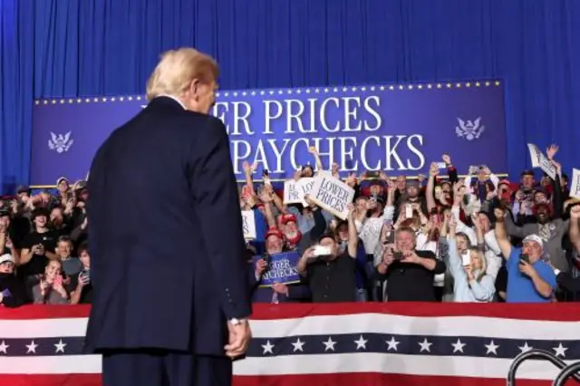 A crowd waves signs as President Trump looks back at them with his arms by his sides