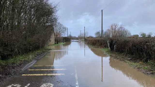A flooded country road flanked by a hedgerow on one side and trees and a house on another.