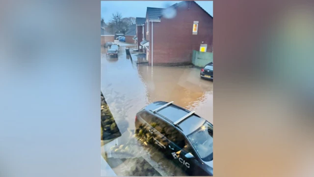 A suburban new build estate covered in floodwater. There are a number of stranded cars.