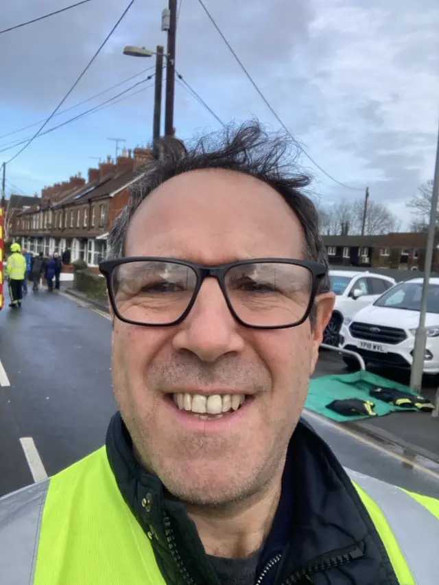 Matt Faulkner smiling into the camera. Emergency services are behind him, dealing with floodwater.