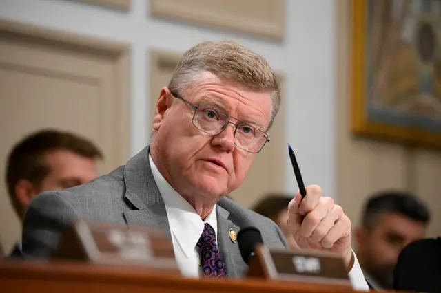 Mark Amodei seated and holding a pen. He is wearing a grey suit over a white shirt and purple tie. He is also wearing eye glasses.