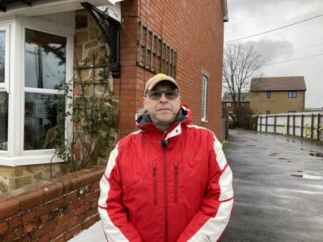 Brian Hamilton wears a read and white waterproof jacket, brown baseball cap and glasses. He is standing in front of a red brick building and looking straight at the camera.