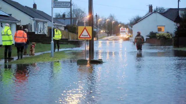 People stand looking at a flooded road near Taunton in Somerset. The photo is taken just after the dawn and the street lights are reflected in the water