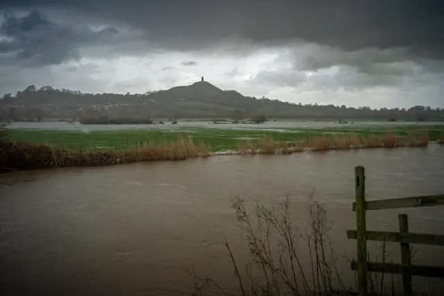 Large dark clouds loom over Glastonbury Tor in the background. In the foreground there is a very swollen river and waterlogged fields.