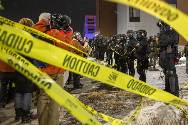 Line of protesters and ICE agents outside a hotel, yellow caution tape in the foreground.