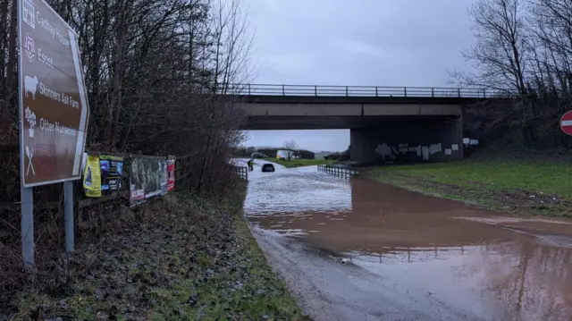An underpass in Devon. There is brown floodwater on the ground. The sky is grey and filled with clouds.