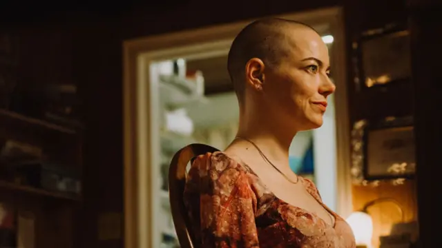 Emma Stone in character in Bugonia - she has a shaved head and wears a flowery dress and gold chain - she is sitting in a room, in a wooden chair