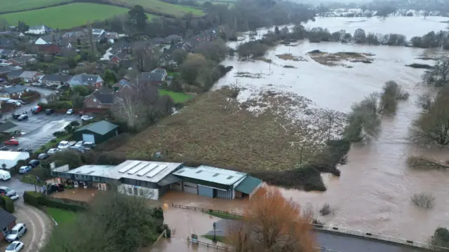A field that has flooded. In front of the field is a building which appears to be flooded.