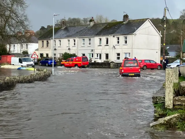 Red emergency vehicles driving through a flooded road. There are houses in the background.