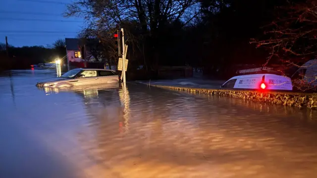 A 4x4 type vehicle stranded in flood water in the dawn light. The water reaches almost to its windows, while a wall behind is almost submerged.