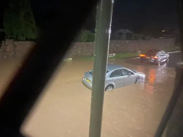 A car submerged in water on the road. There is a black car out the other end.
