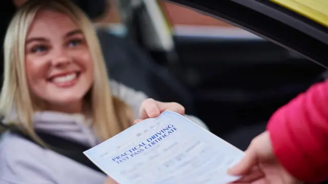 A young woman with blonde hair. She is sat in a car with the seatbelt on. Someone is passing a driving test pass certificate through her open window.