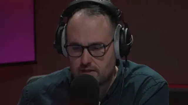 A man in a radio studio with headphones on speaking close to a microphone. He is wearing a blue-green shirt and has dark-rimmed glasses on. In the background is the corner of a TV monitor on a wall.