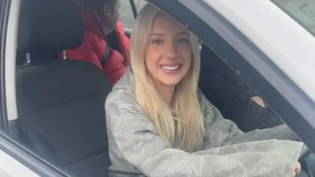 Ira has blonde hair and she is sitting in a car, at the wheel. She is smiling and wearing a grey hoodie. There is someone beside her and in the backseat.