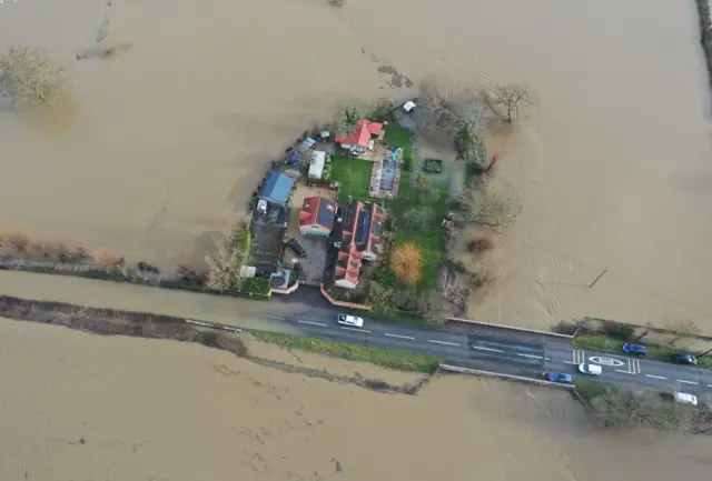 An aerial view of a small cluster of houses completely surrounded by water except for a short stretch of road that is still above the water line.