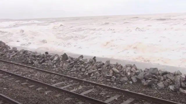 Dawlish sea wall broken. There are rail tracks in front of the wall. Behind the wall are sea waves.