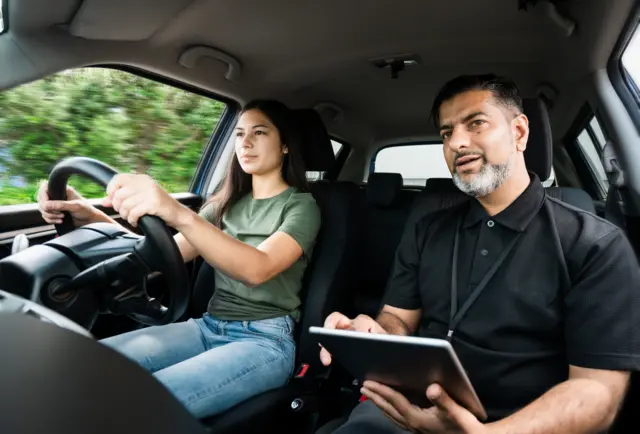 A young woman driving a car with a male driving instructor in the passenger seat. She has long, dark hair and is wearing jeans and a green t-shirt. The man has short, dark hair and a grey beard. He is wearing a black polo shirt and is holding a notebook