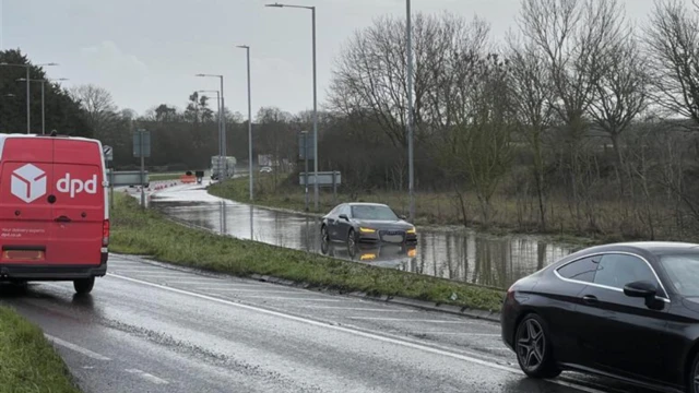 Cars on a busy A-road on a wet day. One saloon-type car is driving through deep standing water on the road.