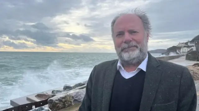 Brazil is wearing a grey jacket over a blue jumper and white shirt and is standing next to a stormy coastline