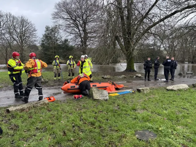 People in high visibility clothing and helmets and people in police uniform. There is grass, in the background there appears to be flooding. There is an orange lifeboat.