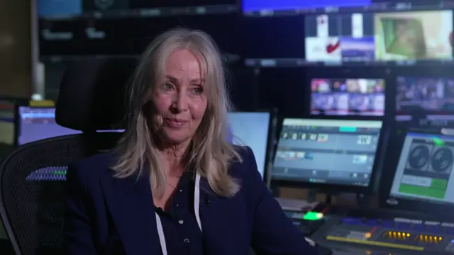 Julie Anne Bailie, a woman with shoulder-length straight blonde hair, smiles during a TV interview. She is stiting in a TV studio gallery with multiple monitors and a mixing desk behind her.