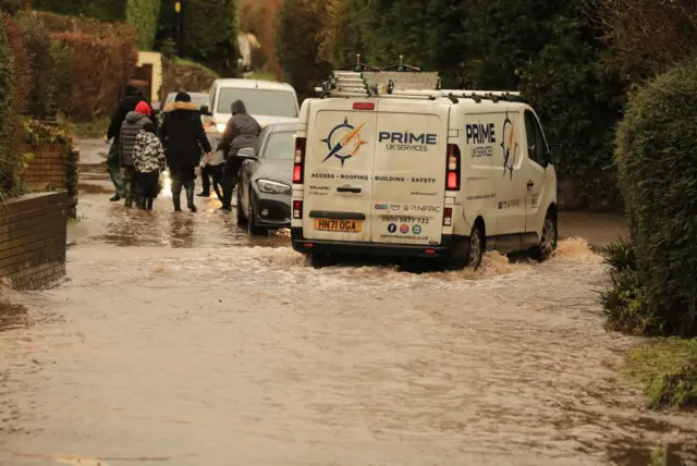 Cars try to navigate flooded roads in a village. A white van appears to be stranded. One family is crossing the floodwater on foot.