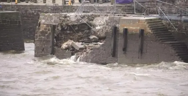 The stone wall of a pier showing damage and falling barriers