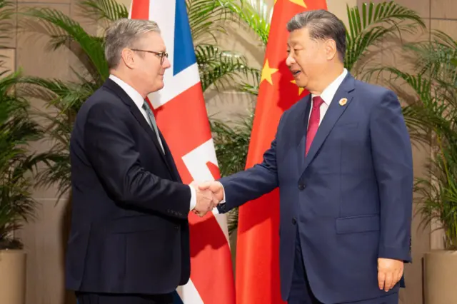 Keir Starmer (L) in black suit shakes hands with Chinese President Xi Jinping (R), in a dark blue suit. They're posing in front of a Union flag and Chinese flag
