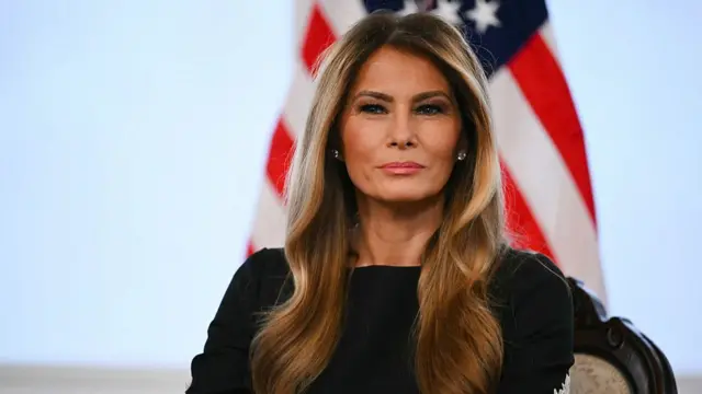 Melania Trump in a black dress with white lace details on the arm, she is sat in front of an American flag.