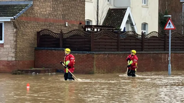 Two firefighters with red uniforms and yellow helmets wade through brown floodwater in Ilminster in Somerset