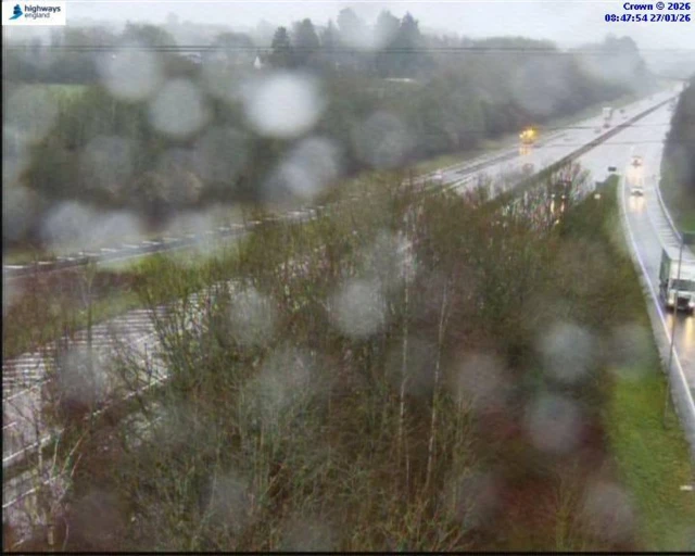 The junction of a major A-road on a rainy day. There are a number of vehicles on the road. Surface water is visible.