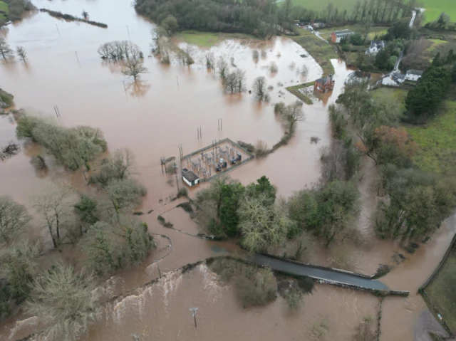 A electricity substation in Devon submerged in flood water, as well as its surroundings. There are trees around it and the station at the centre.