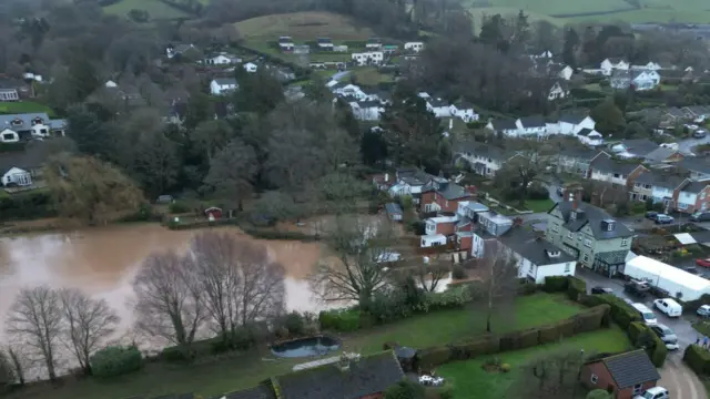 A small town, there are green fields. There is a large amount of brown flood water.