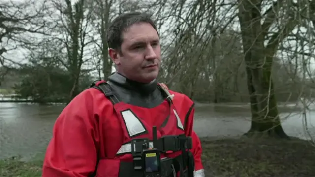 A man in red flood gear and a red life jacket. Behind him is floodwater and several trees.