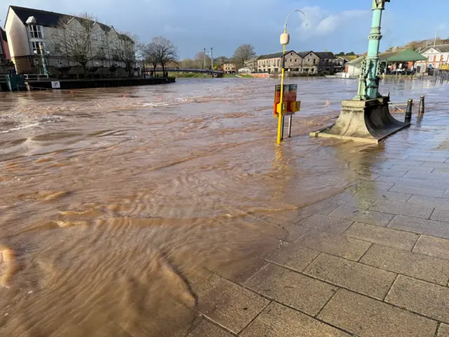 A pavement with a lamp post. There is flood wanted on the pavement and around the blue lamp post. The sky is blue.