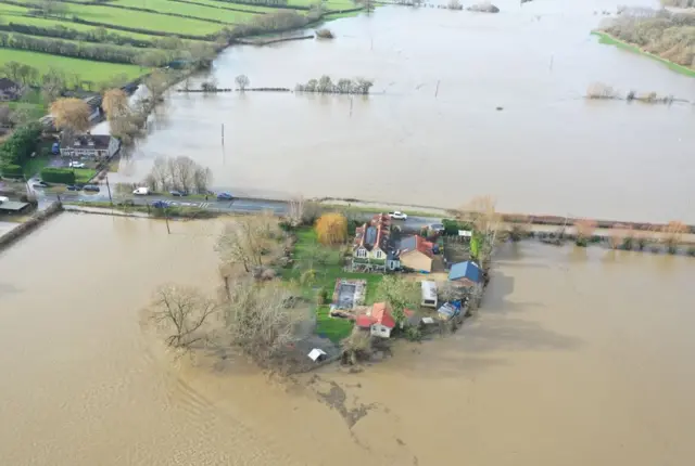 An drone shot of a small cluster of houses completely surrounded by water except for a short stretch of road that is still above the water line.