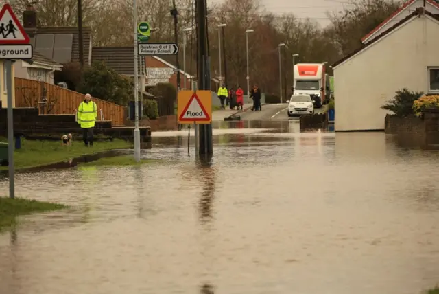 Deep floodwater on a road in a rural village. A queue of cars is visible on the other side, while a man in a high-vis jacket walks his dog. A sign says Ruishton C of E primary school.