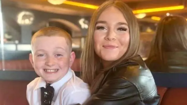 A young boy and teenage girl are sitting side beside - the girl on the right has her arms around the boy and both are smiling widely to the camera. The girl has shoulder length blonde hair and is wearing a black leather jacket. The bot has very short, shaved hair and is wearing a white shirt and tie, with a pair of sunglasses hanging from the front of the shirt's collar. They are sitting on a leather long sofa, topped with mirrors.