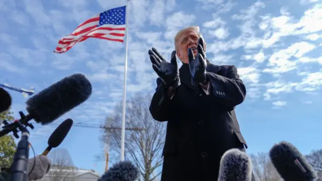 Donald Trump speaking in front of microphones outside the White House with an American flag flying in the background.